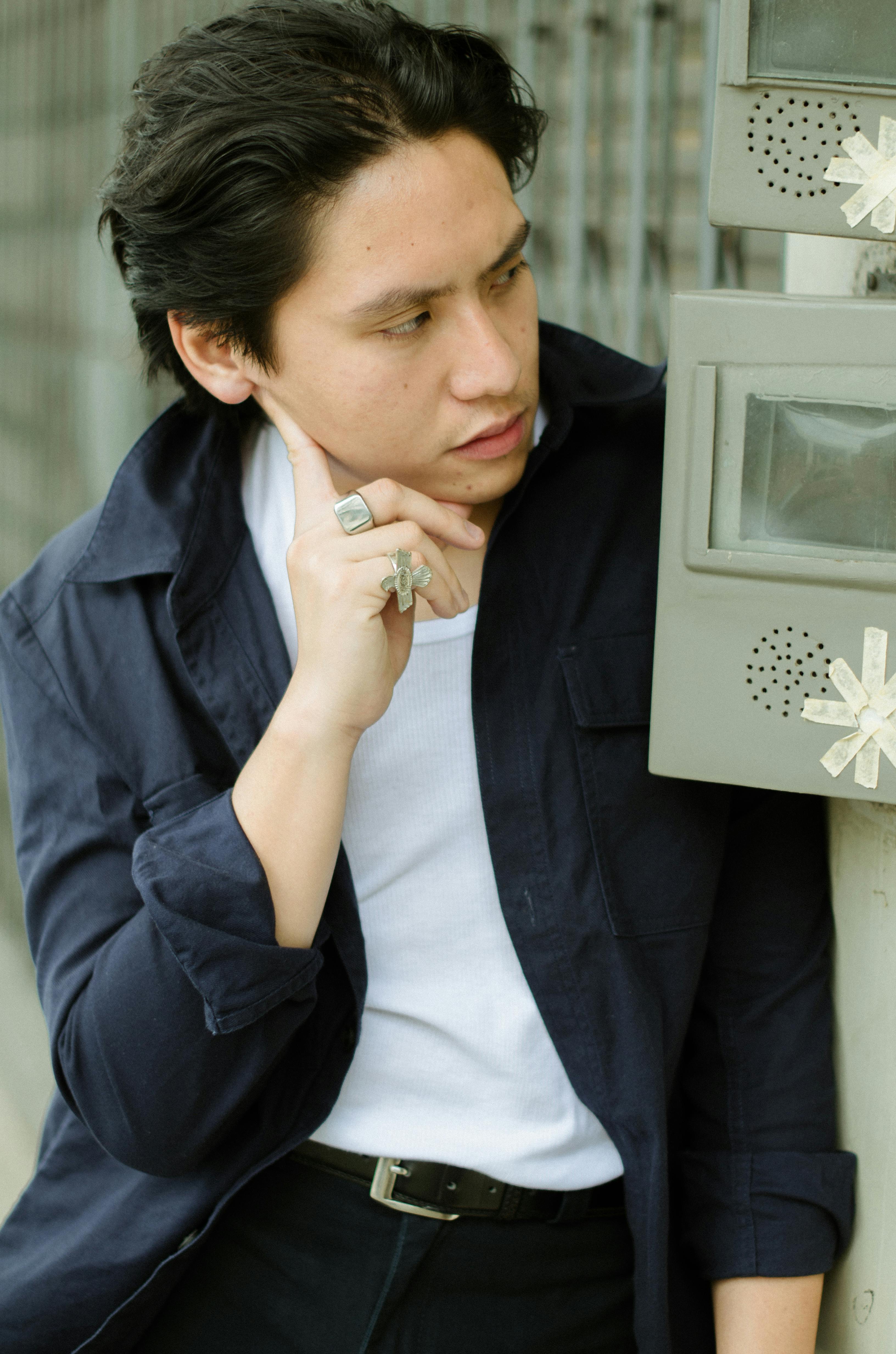 Young stylish ethnic man in jacket touching cheek and looking away standing at metal construction