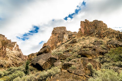 From below of hill slope with rough rocky formations and small bushes under cloudy sky in daylight