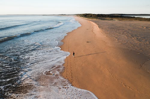 From above of distant traveler walking along sandy coastline near foamy sea on summer day in tropical country in nature