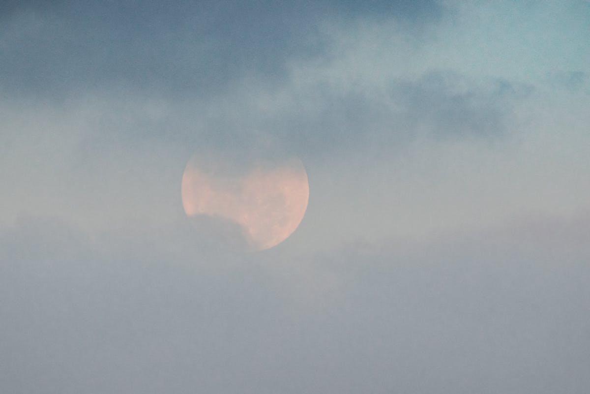 White moon showing through thick clouds on blue sky in gloomy weather on summer day in nature high in atmosphere