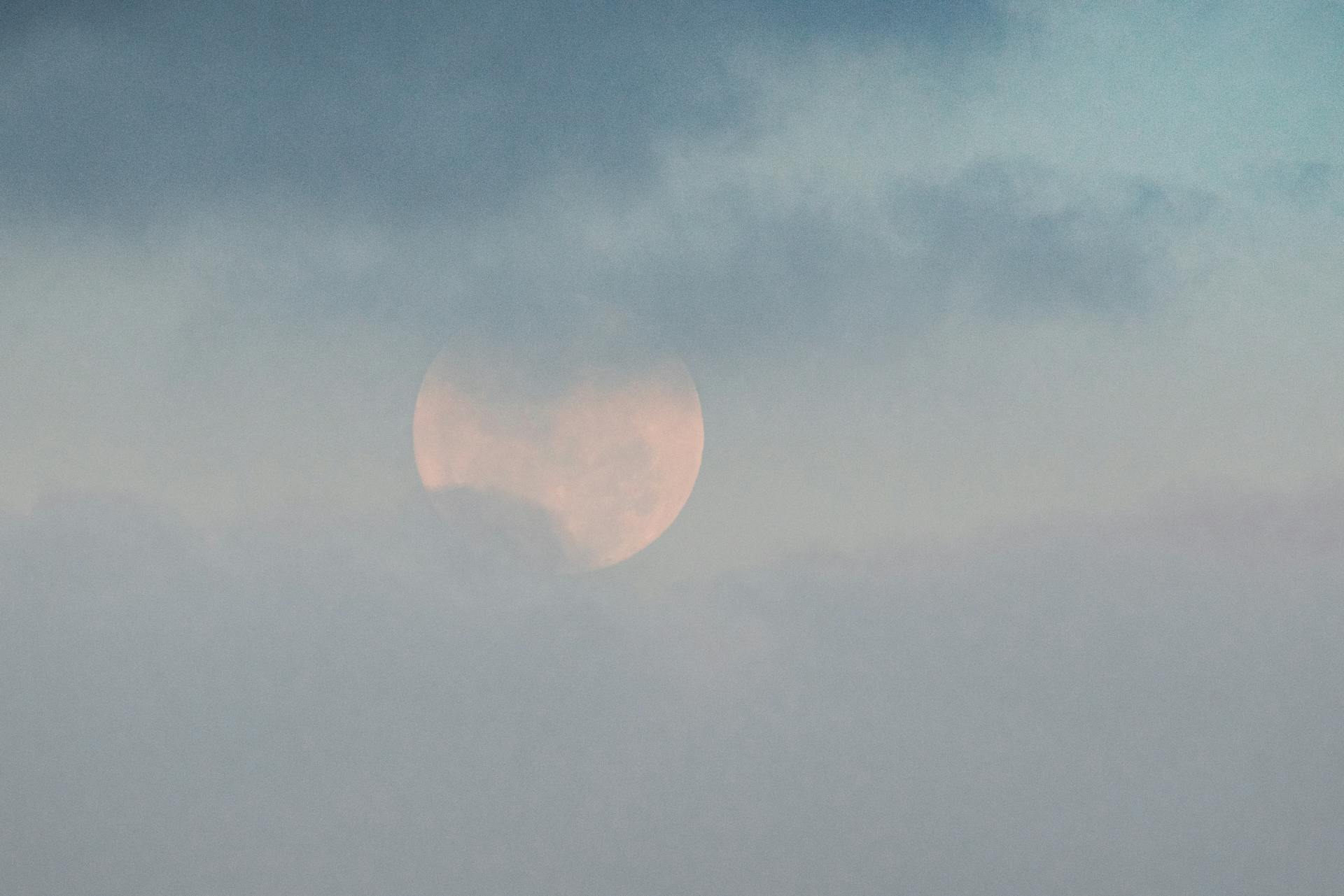 White moon showing through thick clouds on blue sky in gloomy weather on summer day in nature high in atmosphere