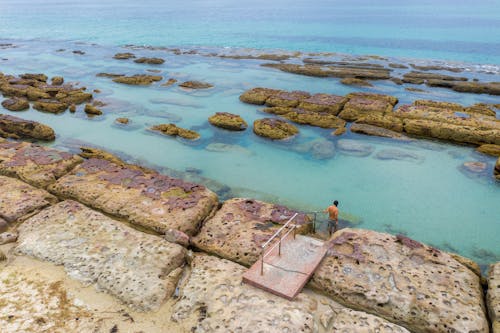 From above of distant traveler standing on metal staircase on stony shore near calm azure water in tropical lagoon in nature