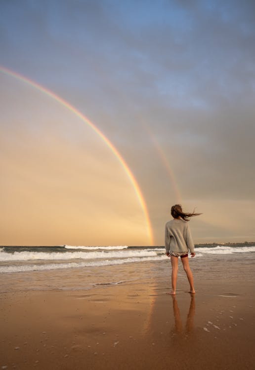 Free Full body of anonymous barefoot female with flying hair standing on sandy coast near waving sea against colorful rainbow in nature Stock Photo