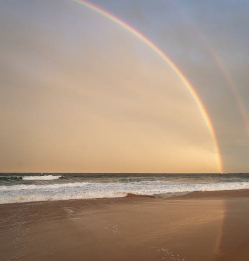 Picturesque scenery of bright rainbow on cloudless sky over waving sea washing sandy coast in nature in tropical country in summer