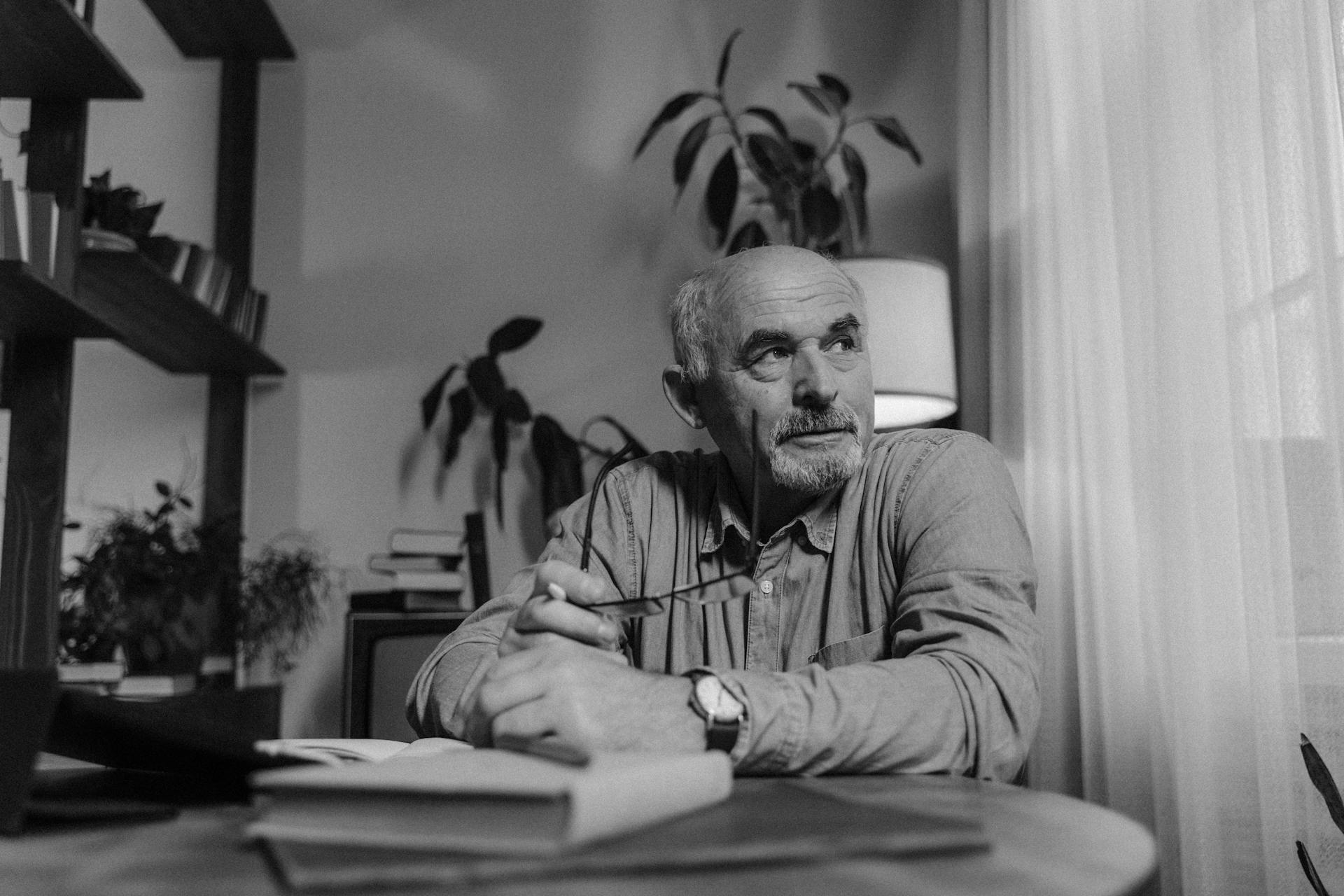 Elderly man sitting in a home office holding eyeglasses, deep in thought. Monochrome image.