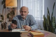 一位老年男子在家中使用智能手机进行网上购物，信用卡放在办公桌旁的植物附近