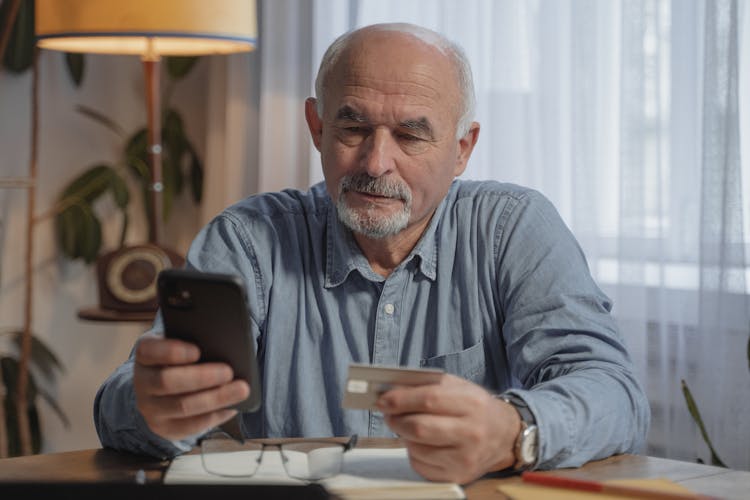 An Elderly Man Holding His Mobile Phone And A Credit Card