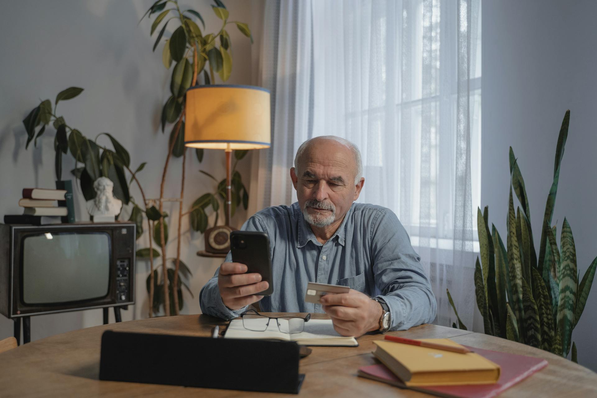 An elderly man sits at a table with a smartphone and credit card, making an online purchase.