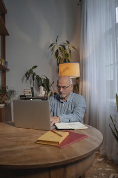 Elderly man with eyeglasses working on a laptop at a wooden table in a cozy home office setting.