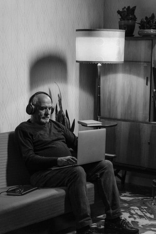 A Grayscale Photo of an Elderly Man Sitting on the Couch while Using His Laptop