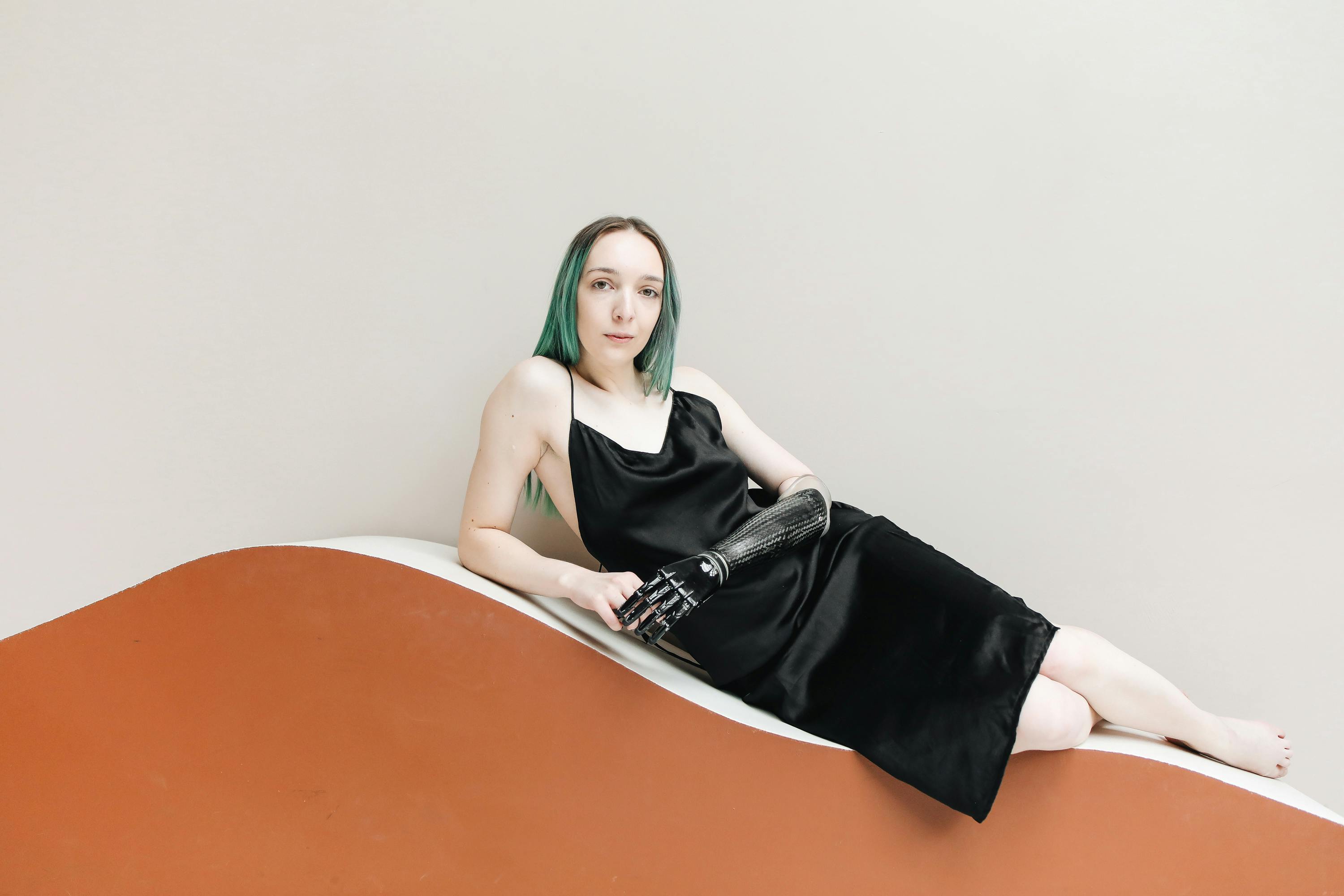 A confident woman with a prosthetic arm posing elegantly in a black dress against a minimalist background.