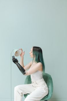 A woman with a prosthetic arm sits on a green chair, adjusting a lamp in a minimalist setting.