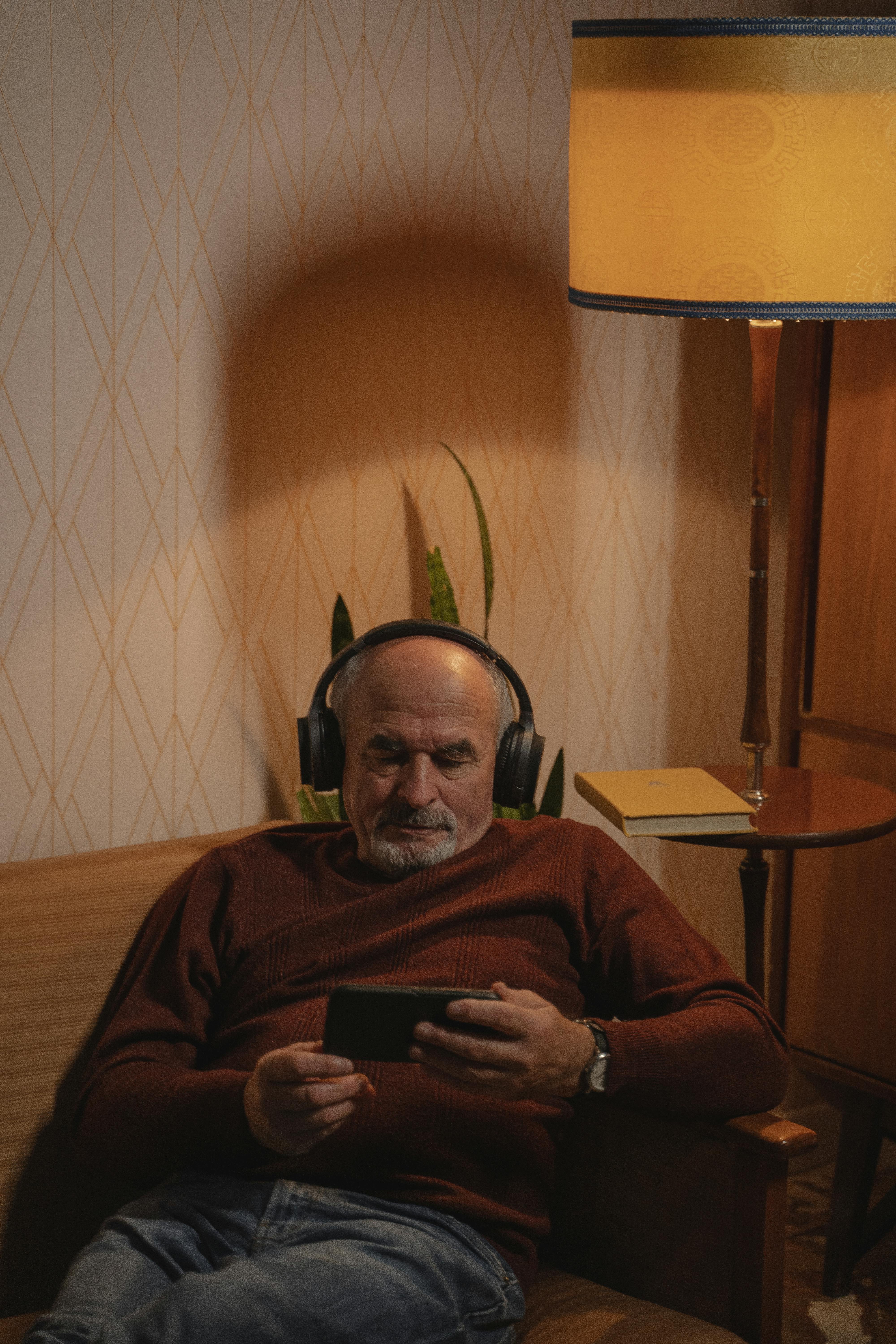An elderly man in a brown sweater enjoys tunes through headphones in a cozy living room.