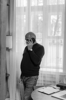 Elderly man talking on cellphone by a window in home office, grayscale image.