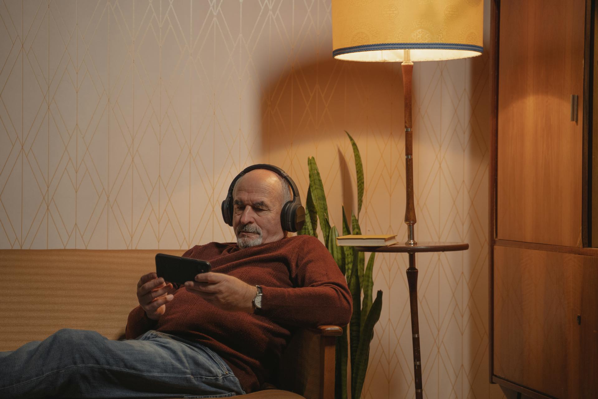 An Elderly Man with Headphones Using a Smartphone while Sitting on the Sofa