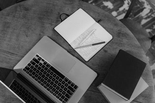 Grayscale Photo of Notebooks beside a Laptop on a Wooden Table