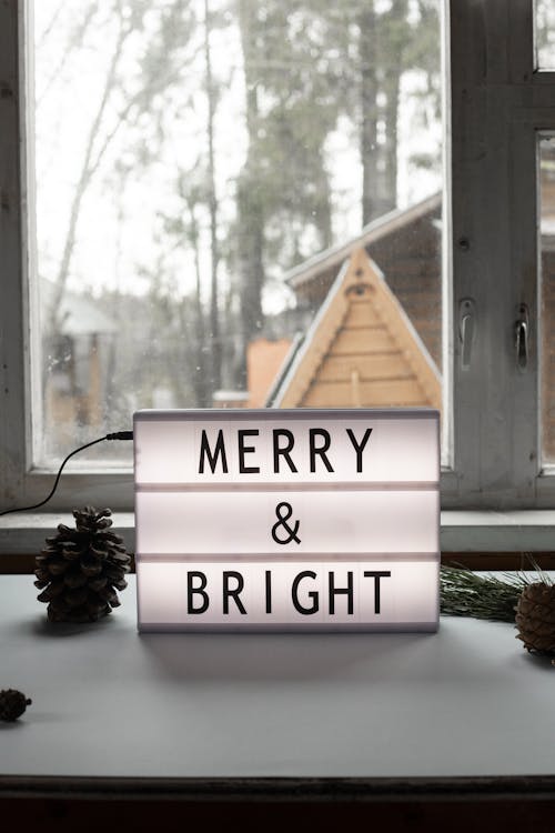 Merry and Bright title on electric signboard on windowsill with pine cones during festive event at home