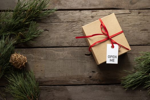 Top view of present cardboard box with ribbon and Open Me title on tag near spruce sprigs on wooden surface during New Year holiday