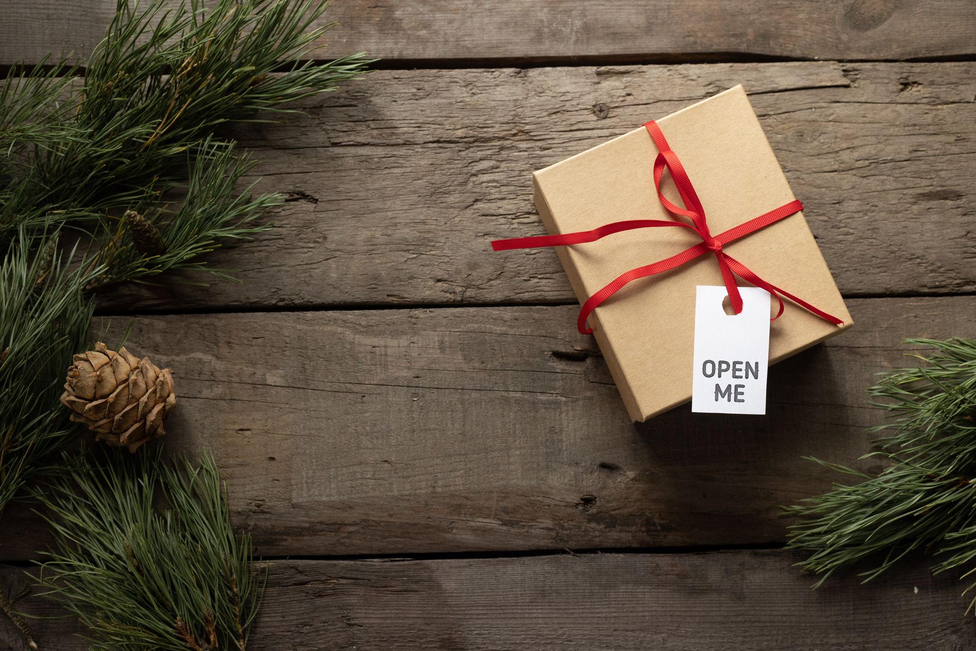 Top view of present cardboard box with ribbon and Open Me title on tag near spruce sprigs on wooden surface during New Year holiday