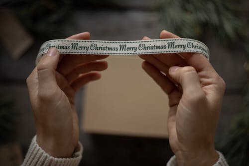 Free Top view crop unrecognizable female showing ribbon with inscription Merry Christmas above gift box during festive season Stock Photo