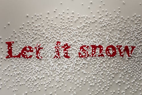 Top view composition of red inscription Let It Snow placed on white surface and covered with artificial snowflakes