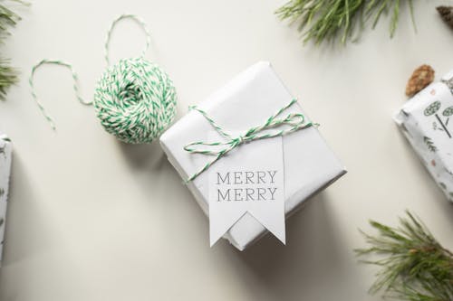 Top view composition of gift box wrapped in white paper with tag Merry Merry placed on table near pine branches and twine during festive holidays