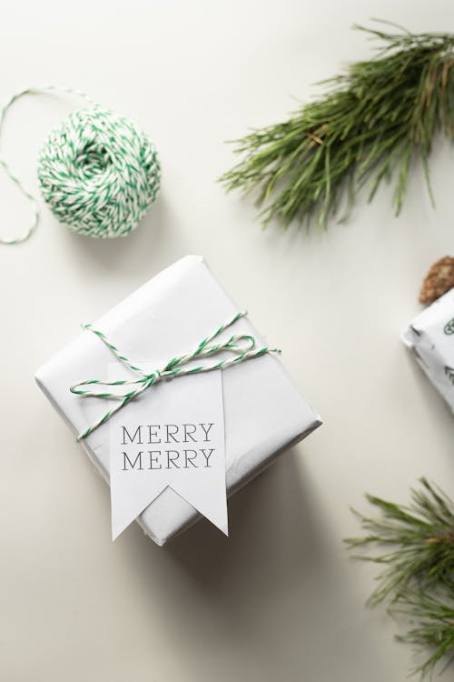 Minimalistic holiday gift wrapped in white paper with a green and white twine and 'Merry Merry' tag, accompanied by pine branches.