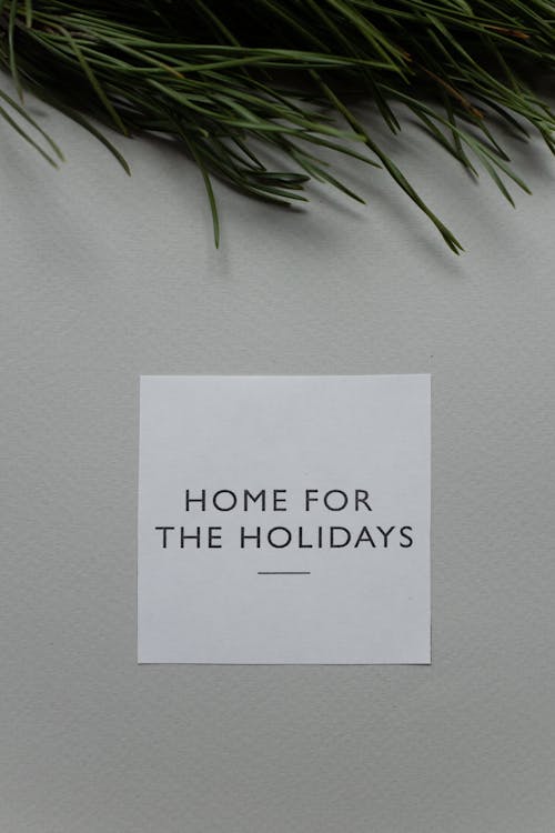 Top view of white greeting card with Home For The Holidays phrase placed on gray background with green coniferous twig