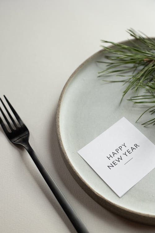 From above of round table with green spruce branch and Happy New Year inscription on card placed on white background with black fork