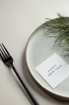 卡片上的铭文置于白色背景上，上方是绿色的云杉树枝和写着“新年快乐”的圆桌，旁边有一把黑色叉子