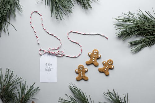 Overhead Christmas composition of gingerbread man cookies placed near gift card with inscription Merry And Bright on white background with green pine tree branches