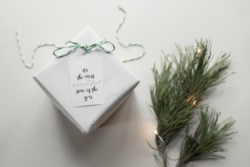 Top view arrangement of wrapped gift box with Its The Most Beautiful Time Of The Year on tag placed on white background near green spruce branches