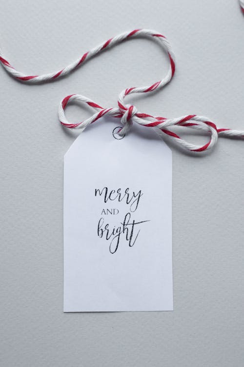 Top view of red and white string tied in bow and attached to white tag with wish Merry And Bright placed on white background