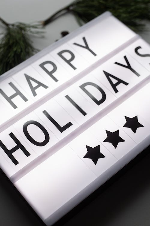 From above of glowing Happy Holidays inscription on white illuminated signboard with stars placed on table with green coniferous branches