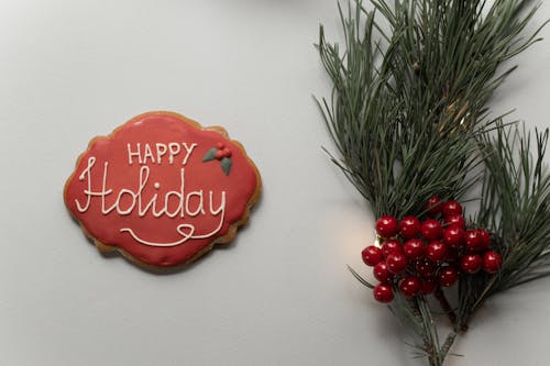 Flat lay composition of gingerbread cookie with red frosting and Happy Holiday inscription placed on gray background with green spruce branch with decorative berries