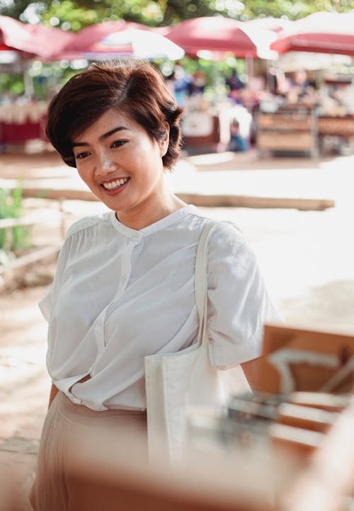 Cheerful young Asian female in stylish white blouse standing on street market on sunny day