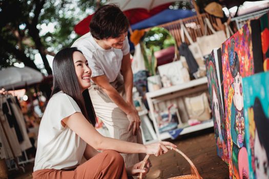 Smiling young Asian females in summer wear looking at vivid colorful drawings in sunny street market