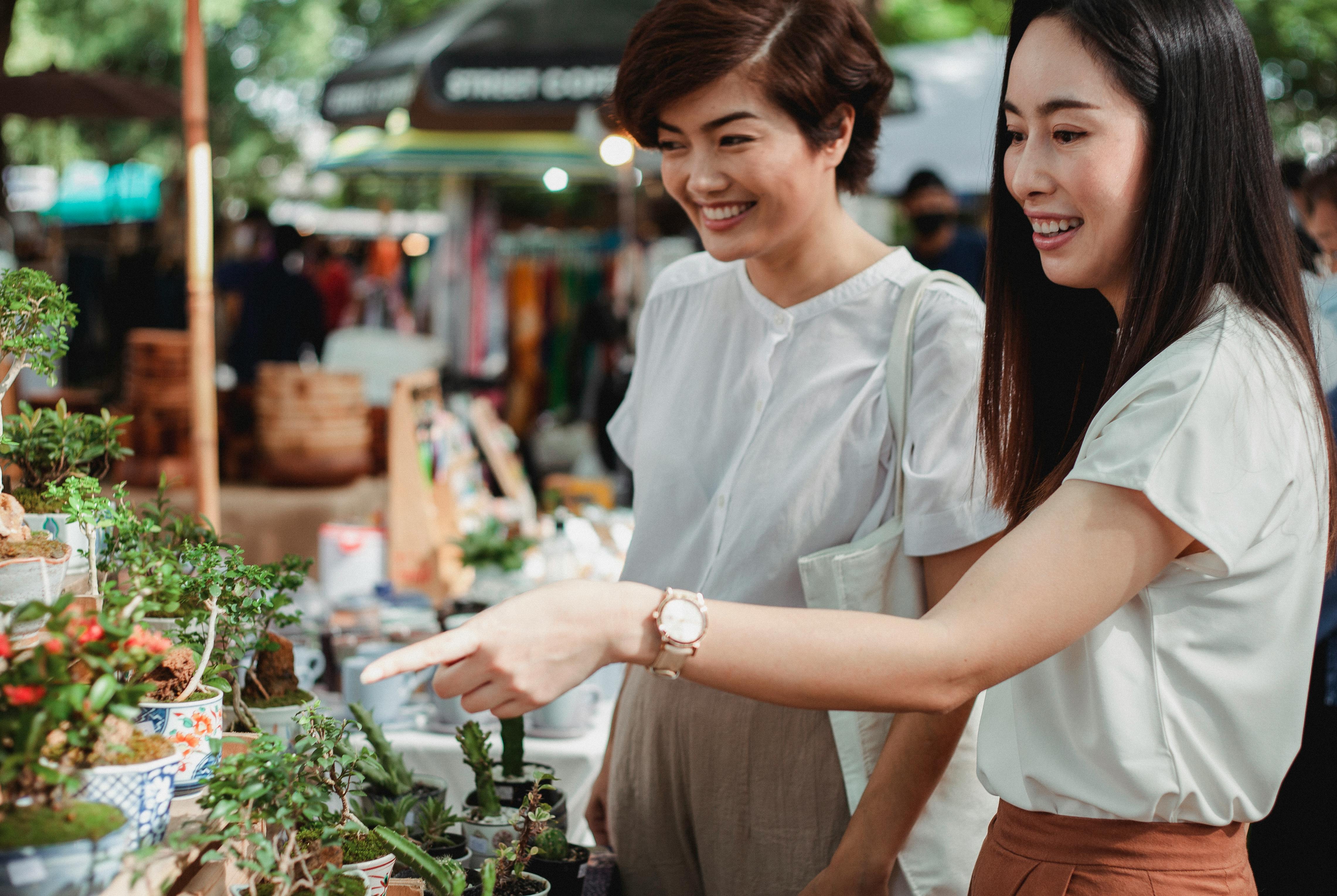 Two women enjoy shopping for plants at an outdoor market, surrounded by greenery and vibrant colors.