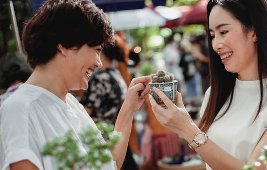 Side view crop happy Asian females in summer outfits looking at young small cactus plant in hand and smiling happily