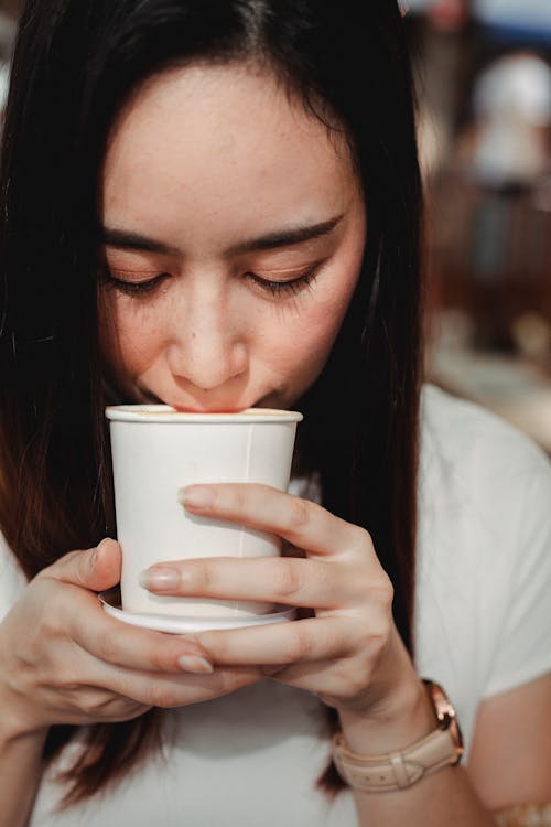 Free Crop serene Asian female wearing casual white shirt drinking with eyes closed aromatic hot drink from takeaway cup Stock Photo