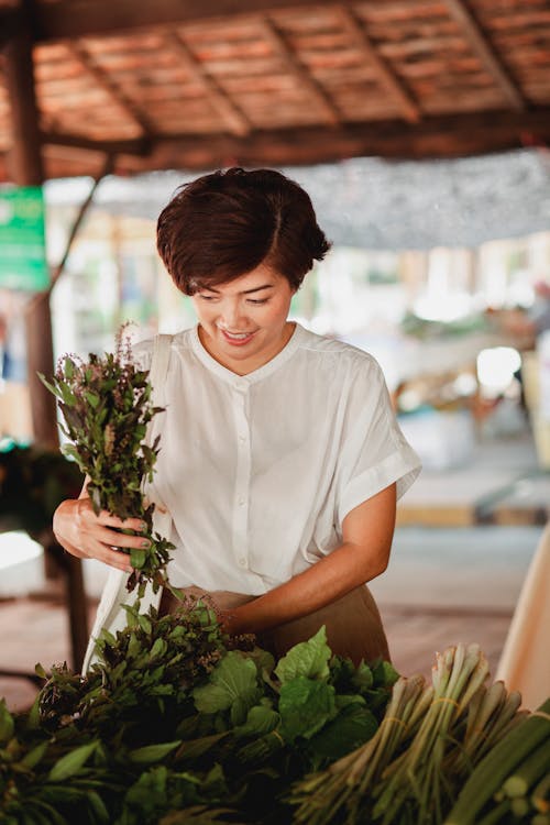 Веселая азиатская женщина, выбирающая свежие травы на рынке