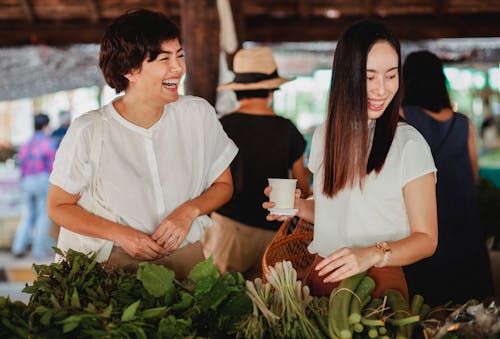 Cheerful Asian female friends picking fresh exotic greenery at oriental street store while laughing together