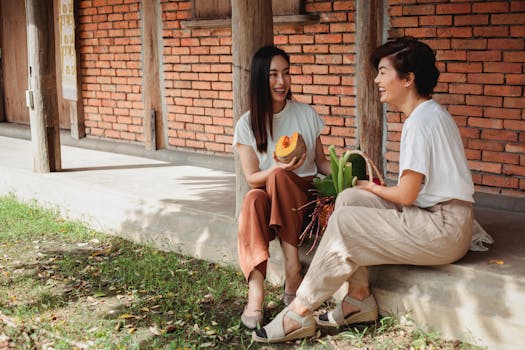 Young laughing Asian girlfriends with basket full of fresh vegetables speaking on street near brick wall