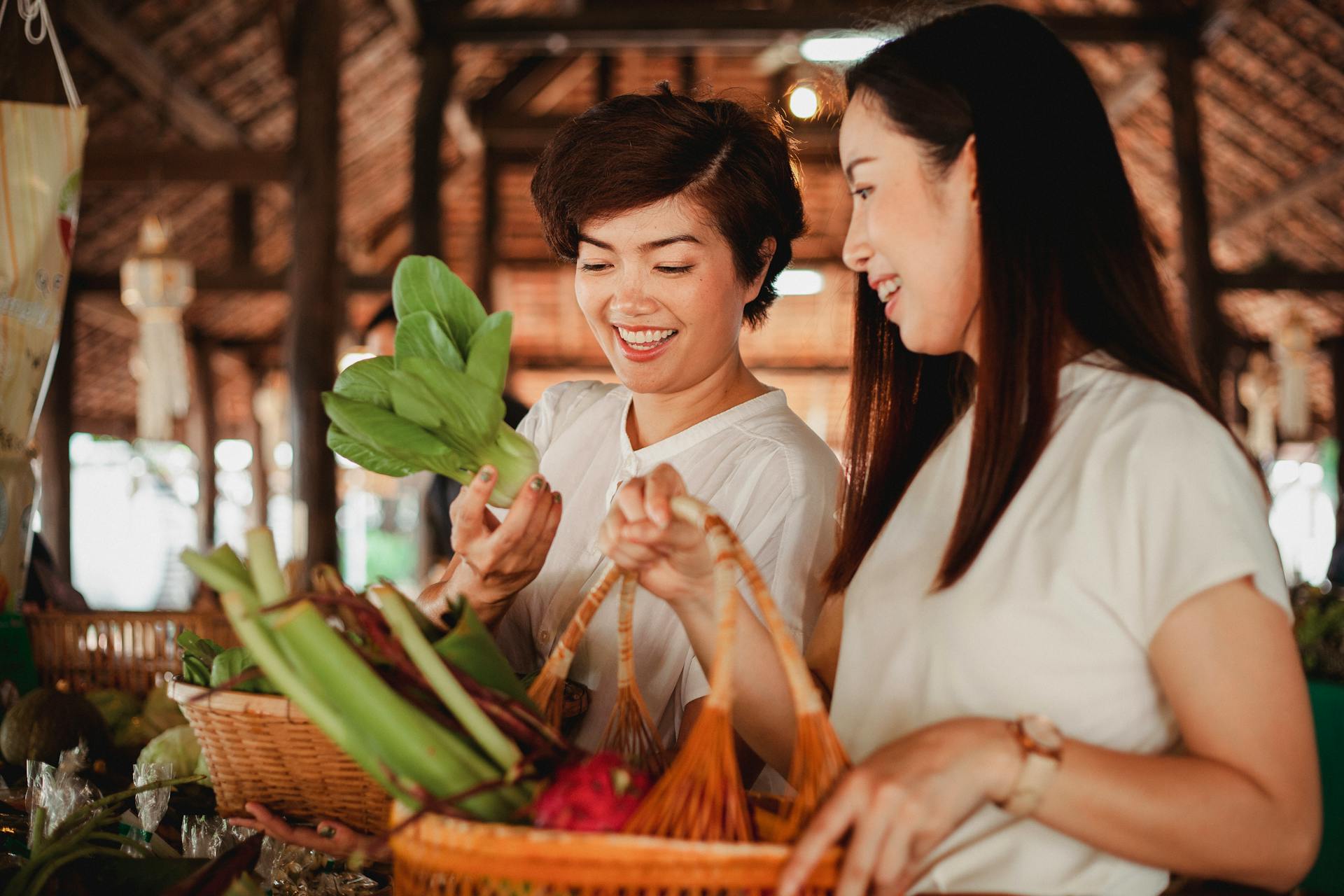 Cheerful Asian female buyers with baskets and fresh Chinese cabbage in oriental market in daylight