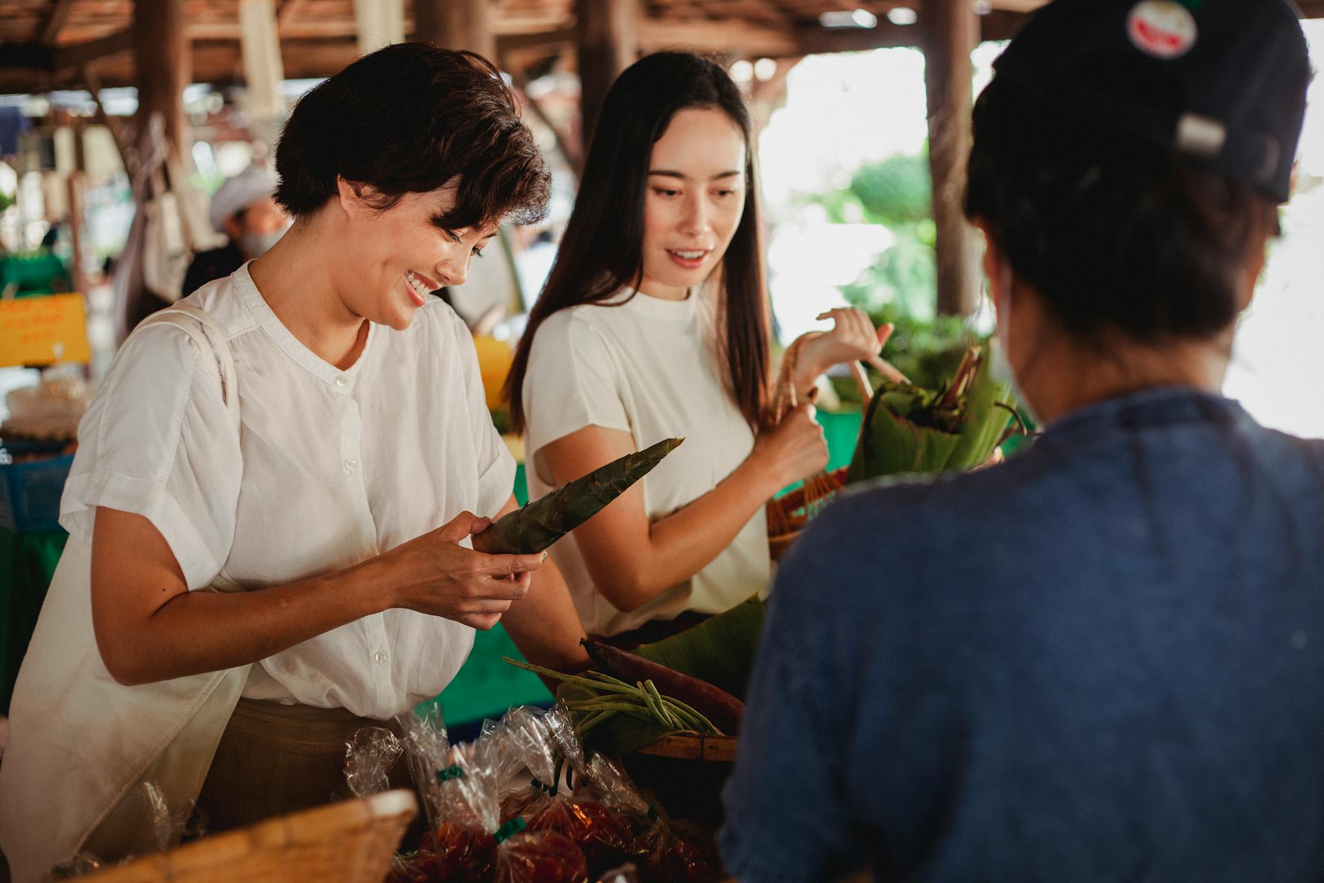 Smiling ethnic shoppers choosing vegetables near unrecognizable seller in market