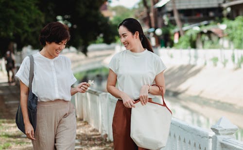 Young smiling ethnic women in casual outfit with bags speaking while strolling on walkway in town