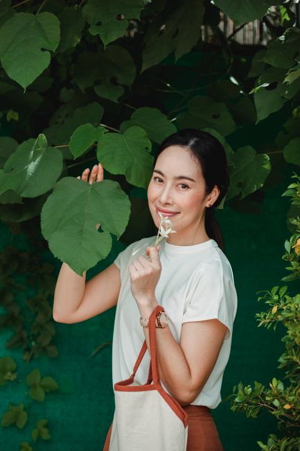 Young sincere ethnic female with flower and bag touching tree leaf while looking at camera in park