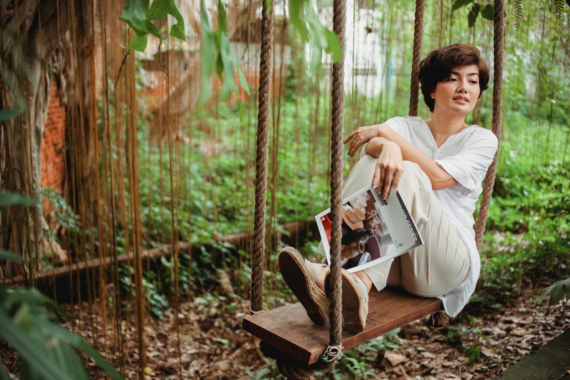 Thoughtful Asian woman with hairstyle in stylish clothes sitting on swing with crossed hands with magazine and looking away