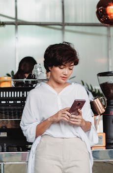 Asian woman at a coffee shop, browsing her smartphone calmly while leaning on the counter.