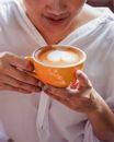 Crop anonymous female in white shirt raising hands with cup of fresh coffee with heart drawn on foam